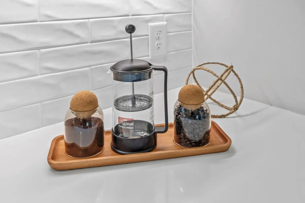 A warmly lit kitchen scene featuring a French press, jars of coffee grounds, and a decorative orb arranged on the countertop. The inviting setup hints at a space where guests can begin their day with freshly brewed coffee or gather for casual conversation.