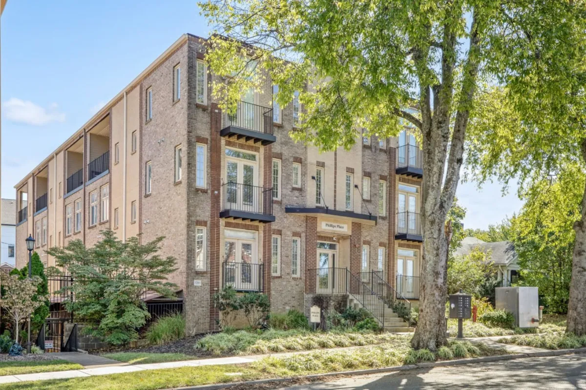 The image showcases the building's welcoming brick exterior, featuring balconies, large windows, and tastefully landscaped greenery. Residents can be seen relaxing on the balconies and socializing in the front yard, all set against a vibrant neighborhood backdrop.