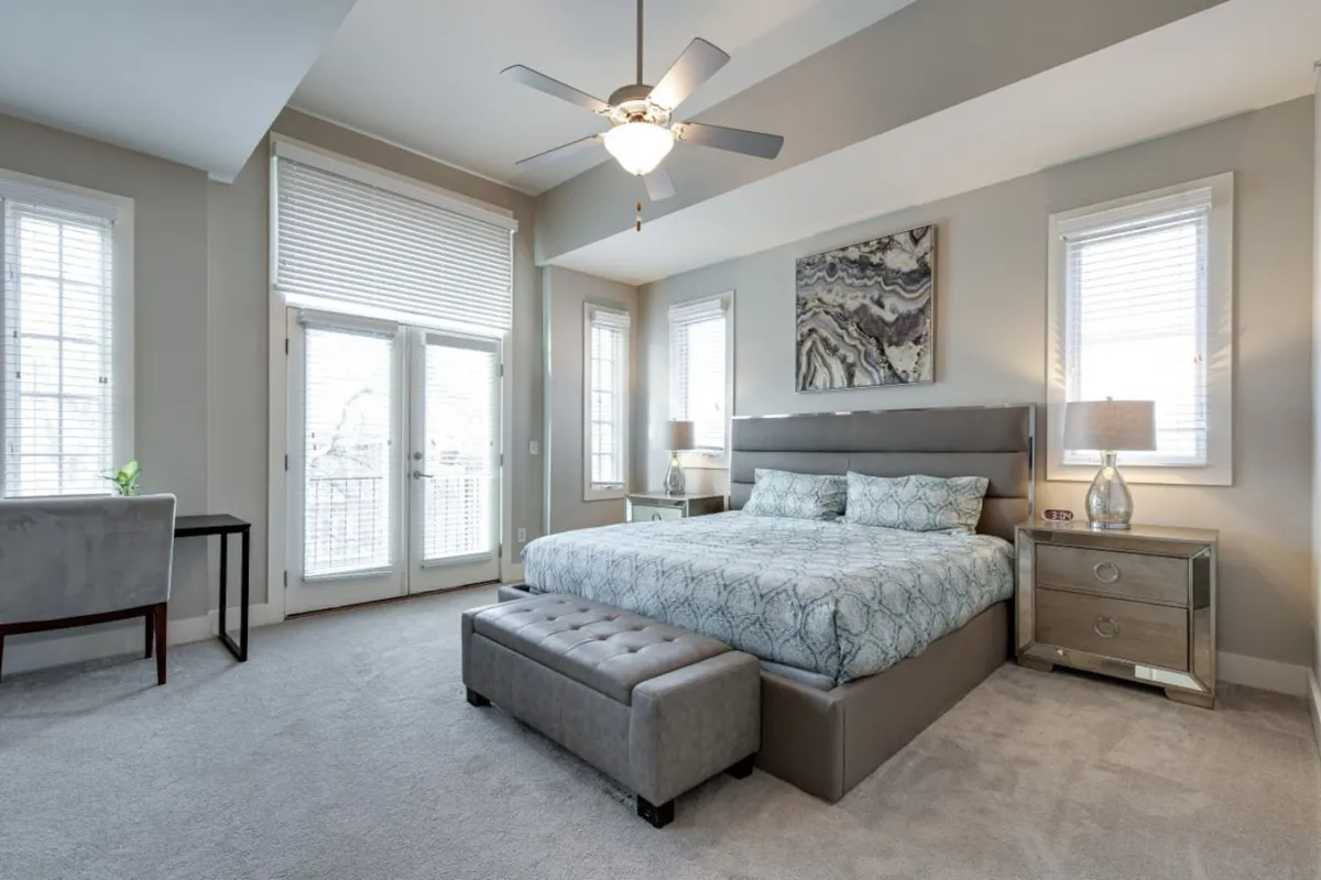 The image showcases a serene and inviting bedroom with a luxurious king bed, stylish nightstands, vibrant artwork above the headboard, large windows flooding the room with natural light, and a cozy seating area—creating a relaxing retreat for guests.