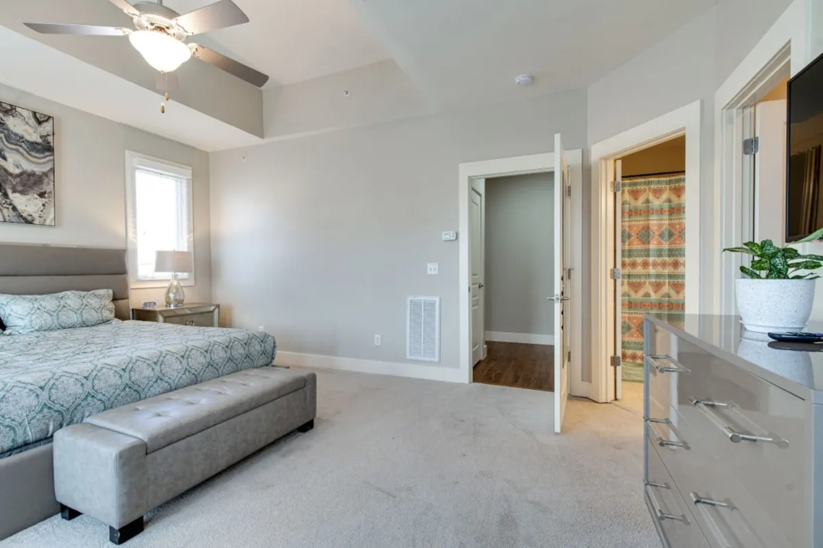 A serene king bedroom featuring decorative pillows on a large bed, a dresser, stylish artwork, and a doorway that leads directly to the ensuite bathroom. The space is bathed in natural light, offering a tranquil spot for rest and privacy.