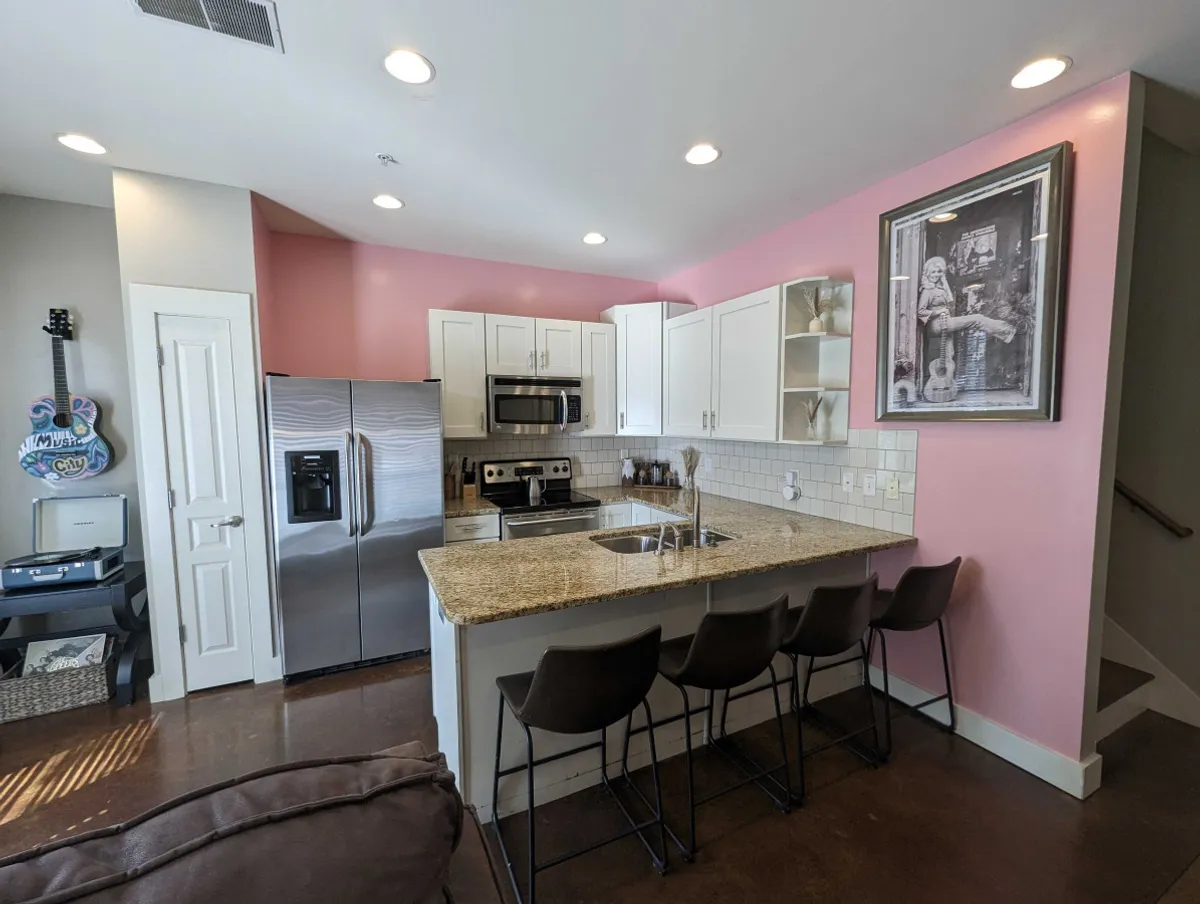 A modern kitchen with a granite countertop, stainless steel appliances, a cheerful pink accent wall, and framed artwork. The space is bright and designed for both cooking and entertaining, making it ideal for brunches, coffee rituals, or toasts before heading out. The setup embodies a fully-equipped kitchen and the dining peninsula described in the section.