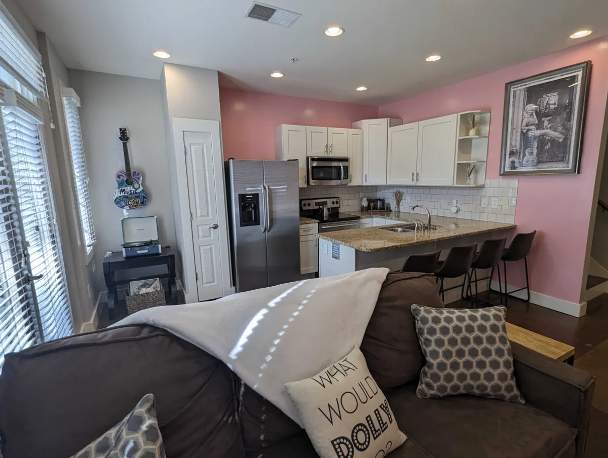 A casual, welcoming interior living space that blends a kitchen island, sofa with decorative pillows, and striking guitar-themed wall art. The room is bright and open, perfect for gathering, relaxing, or getting ready for a night out.