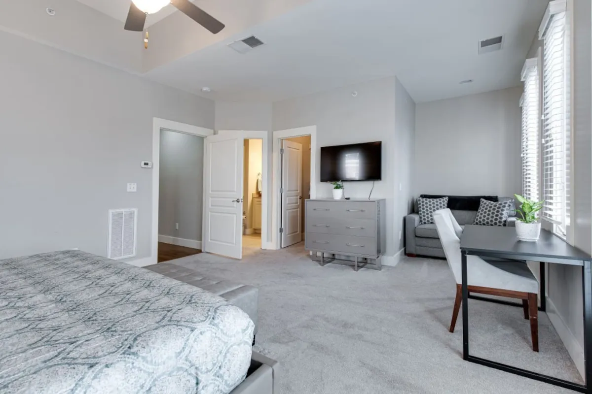 A spacious king bedroom with a large bed dressed in plush bedding, a dresser and TV, a sleek desk setup, and a cozy sofa. Natural light pours in through a window, creating a comfortable and contemporary atmosphere ideal for relaxing, working, or unwinding.