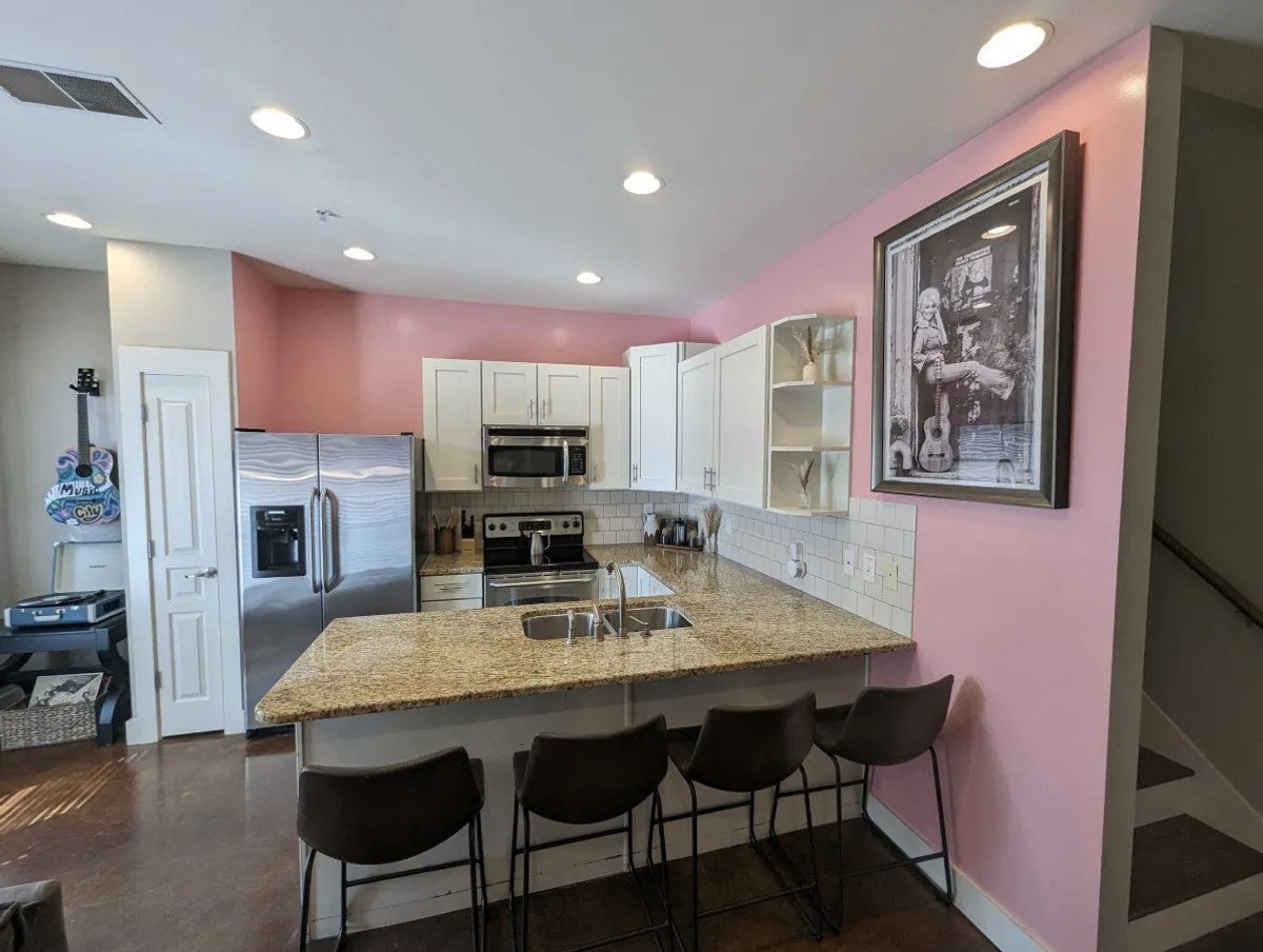 A welcoming kitchen with a spacious island, modern stainless steel appliances, playful pink accent walls, and eye-catching black and white artwork. The space feels inviting, ideal for preparing meals or gathering casually after a workday.