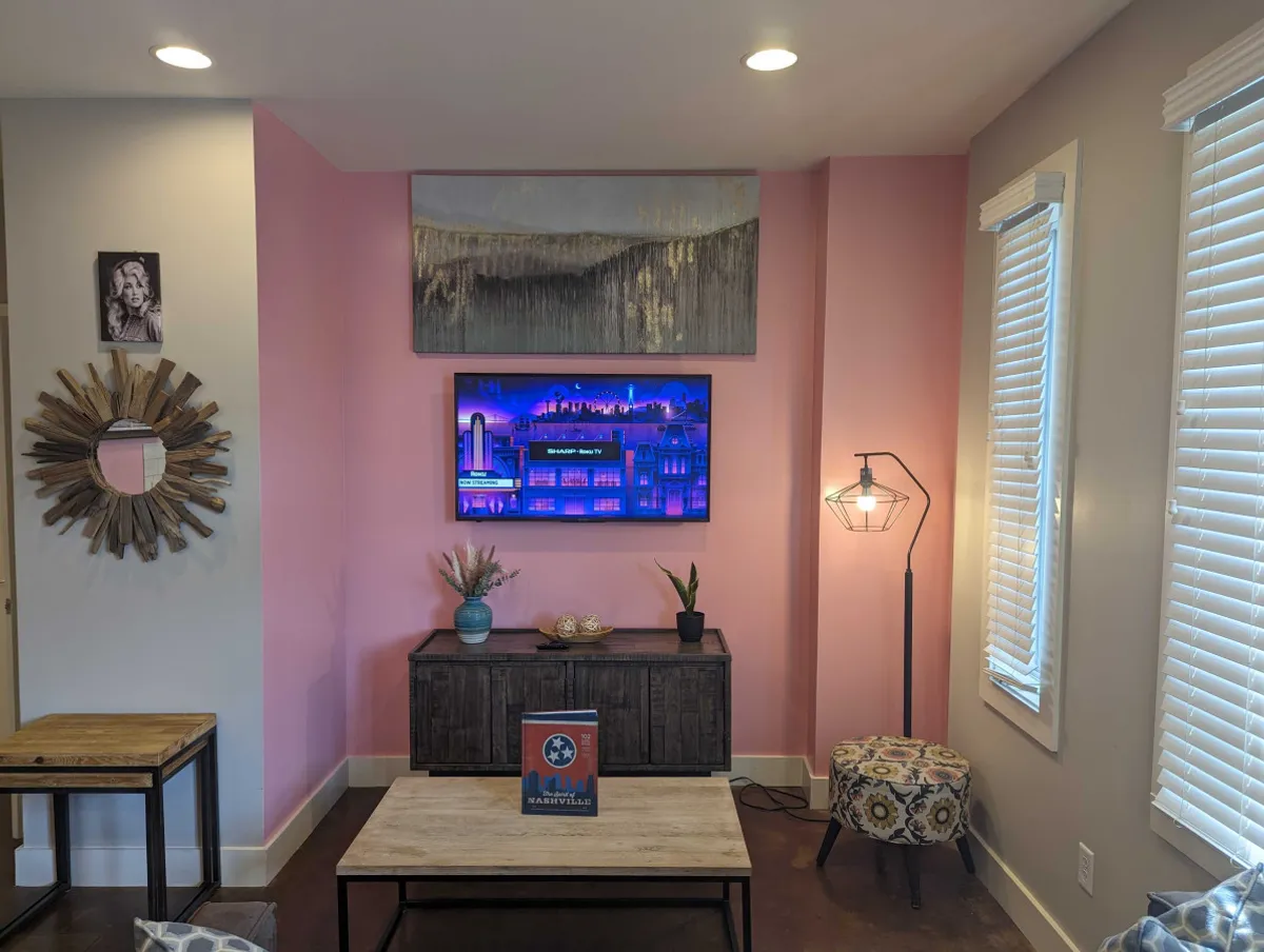 The living space features a smart TV displaying a colorful cityscape, a modern wooden coffee table, a decorative lamp, a sunburst mirror, and a pink accent wall. The arrangement is perfect for relaxing, socializing, or enjoying a movie night with friends or family, enhanced by stylish decor.