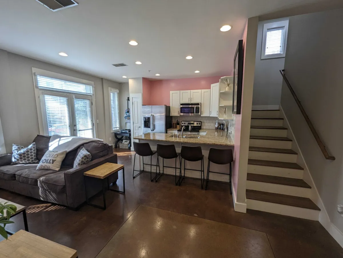 This image captures an inviting living room with a sofa adorned with decorative pillows, a kitchen island with bar stools, and a staircase leading to the upper level. The open-plan layout seamlessly connects the living and kitchen areas, with natural light brightening the space, evoking a sense of warmth and togetherness.